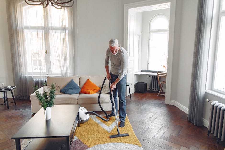 Close-up of a black vacuum cleaner with a rectangular cleaning head, being used to clean a patterned, beige and pastel-colored carpet in a residential space. The vacuum hose extends upward, held by a 