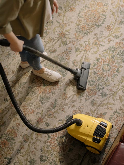 Close-up of a black vacuum cleaner head being used to clean a detailed, patterned area rug with floral motifs on a wooden floor, with a person wearing white sneakers standing nearby. The scene is well