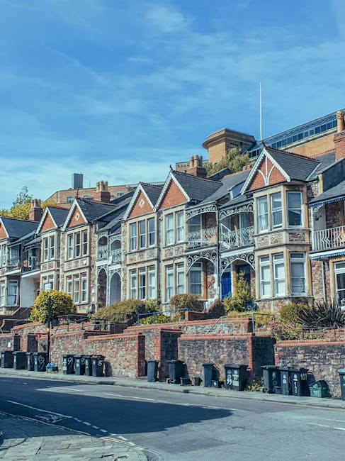 A row of Victorian-style terraced houses with decorative white trim and bay windows, situated on a sloped street under a bright blue sky with wispy clouds. The houses are built with red brick facades,