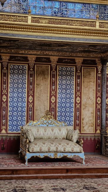 An ornate antique armchair with a blue velvet upholstery and intricate gold wood carving details on the frame, positioned on a lush green artificial turf surface in an indoor sports arena. The chair's